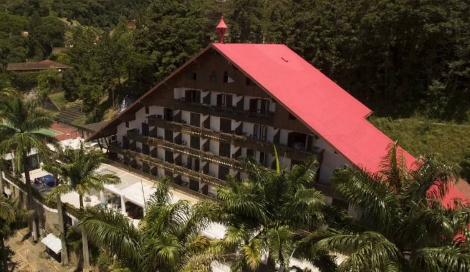 Vista aérea de um grande hotel com telhado vermelho vibrante e inclinado, estilo alpino, cercado por densa vegetação verde. O edifício tem várias varandas com grades de madeira escura, e palmeiras altas em primeiro plano. Ao fundo, mais árvores e colinas.