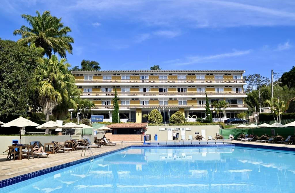 Hotel de cinco andares com fachada amarela e varandas brancas, cercado por palmeiras e vegetação exuberante. Em primeiro plano, uma piscina azul clara com espreguiçadeiras e guarda-sóis brancos, onde algumas pessoas relaxam. O céu azul com nuvens esparsas complementa a cena ensolarada