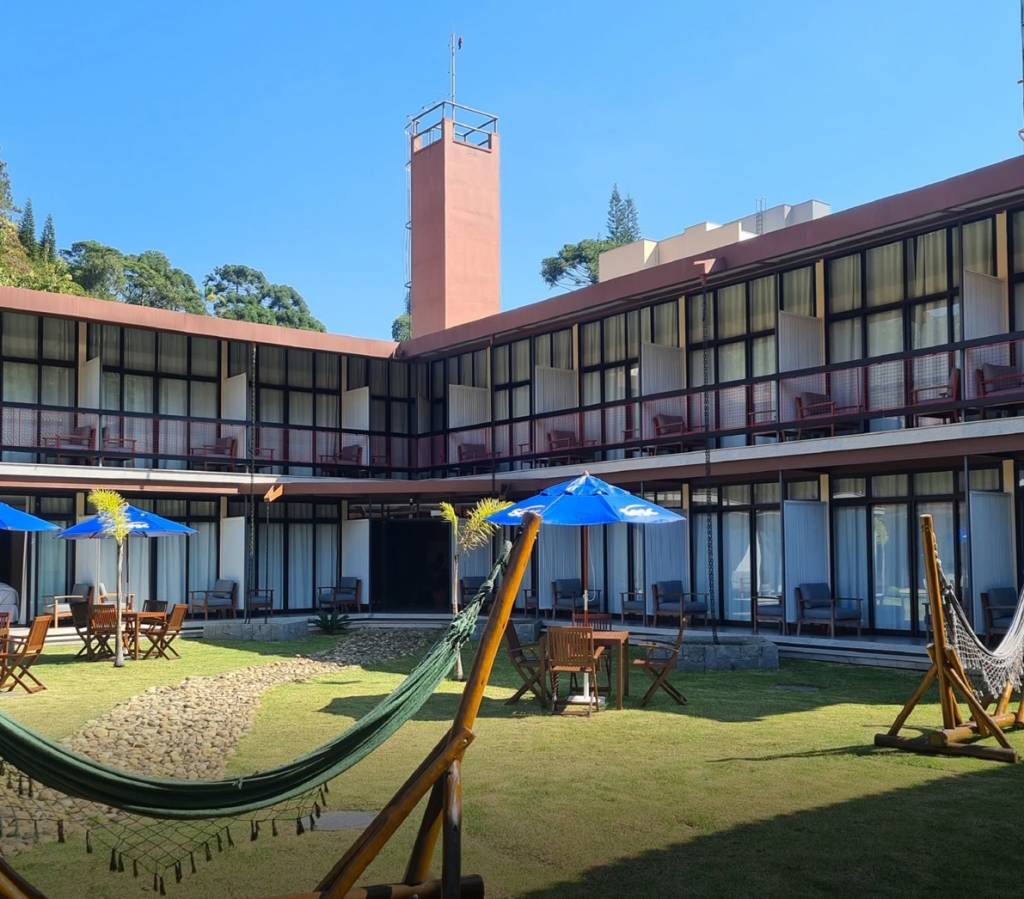 Pátio interno de um hotel com gramado verde, duas redes de descanso, mesas de madeira com cadeiras sob guarda-sóis azuis, e um caminho de pedras. Ao fundo, um prédio de dois andares com varandas e uma torre marrom avermelhada se eleva contra o céu azul
