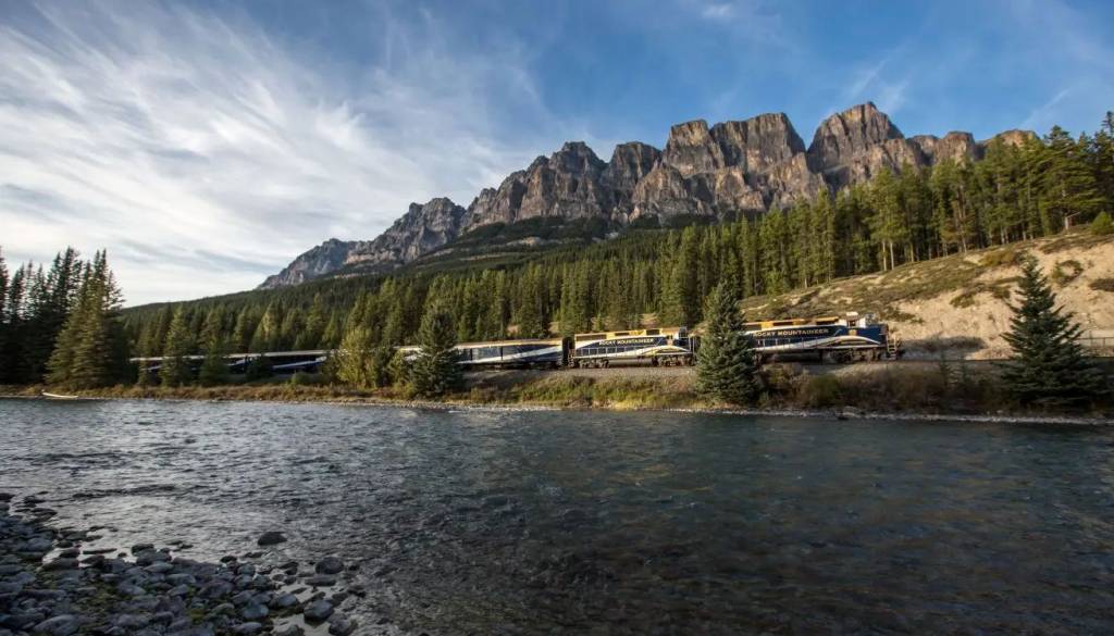 Um trem azul e amarelo com a inscrição Rocky Mountaineer percorre uma ferrovia ao lado de um rio rochoso, com uma floresta de pinheiros e montanhas rochosas ao fundo, sob um céu azul com nuvens esparsas