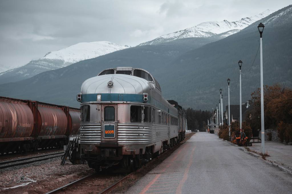 Trem de passageiros prateado com detalhes azuis e o logo VIA parado em uma estação, com vagões de carga marrons à esquerda. Ao fundo, montanhas com picos nevados sob um céu nublado. À direita, postes de luz e árvores com folhagem outonal.