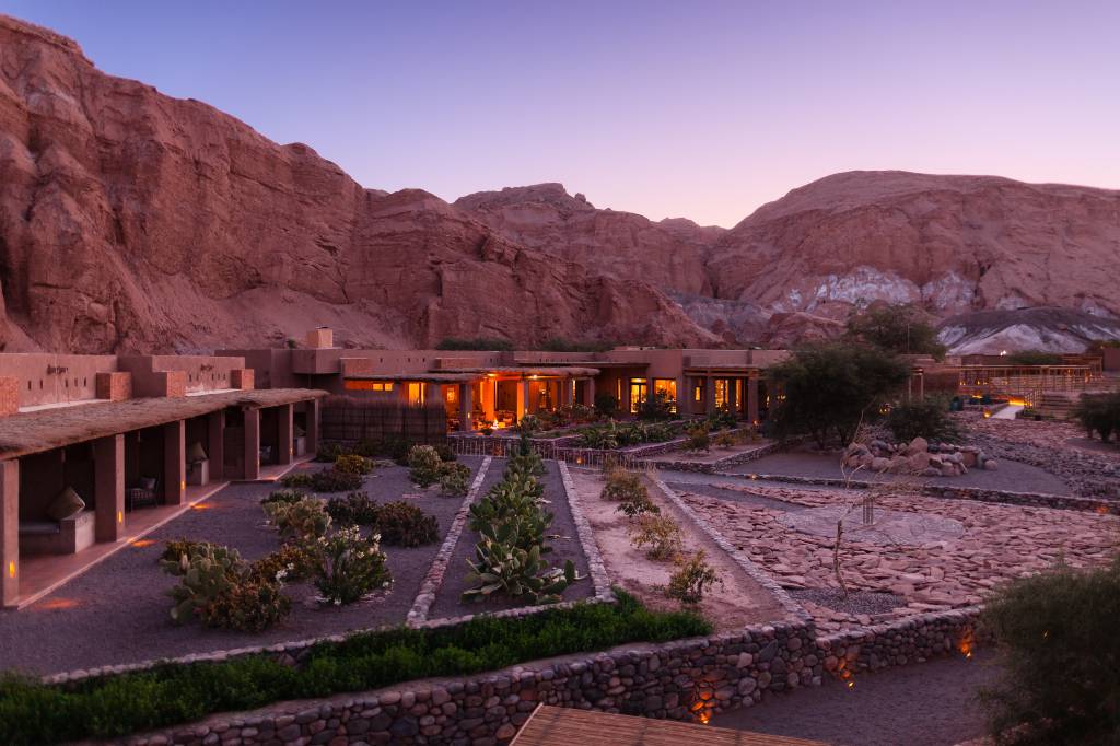 Resort de luxo com arquitetura de adobe em tons terrosos, iluminado por dentro, ao anoitecer. Jardins desérticos com cactos e pedras, e montanhas rochosas avermelhadas ao fundo sob um céu roxo e azul
