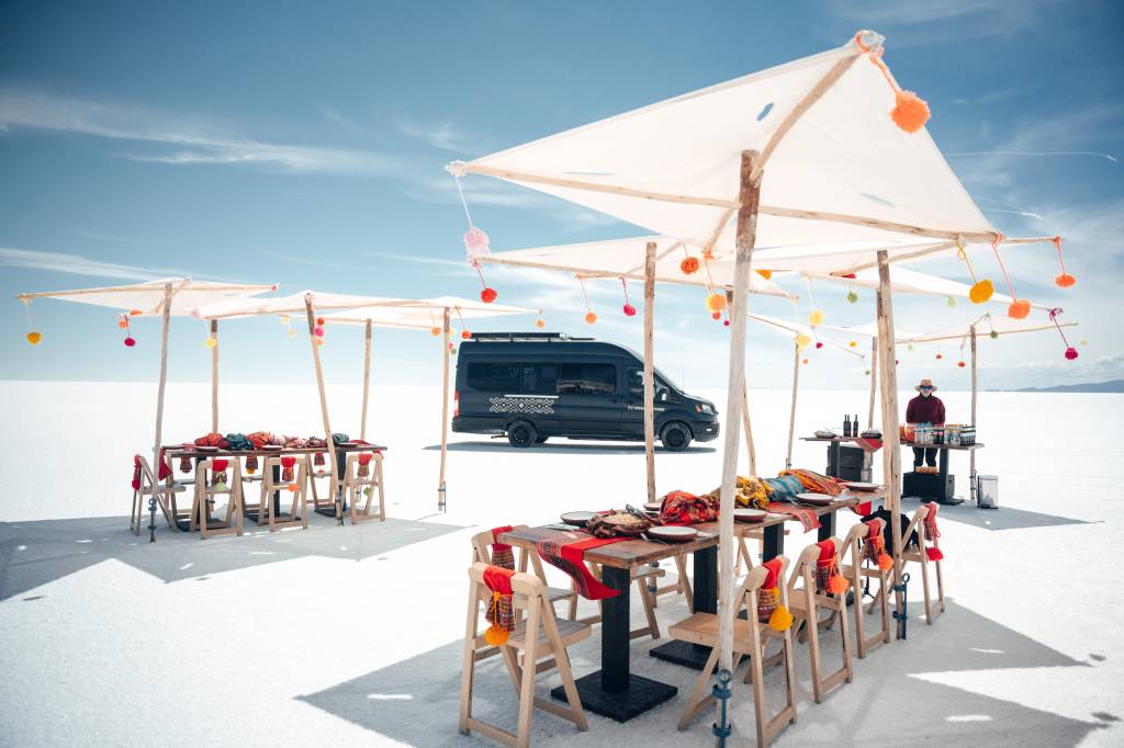 Cenário de refeição ao ar livre no deserto de sal, com mesas de madeira e cadeiras dobráveis sob tendas brancas decoradas com pompons coloridos. Uma van preta está estacionada ao fundo, e um homem de chapéu serve bebidas em uma mesa lateral. O céu azul claro contrasta com o chão branco e plano