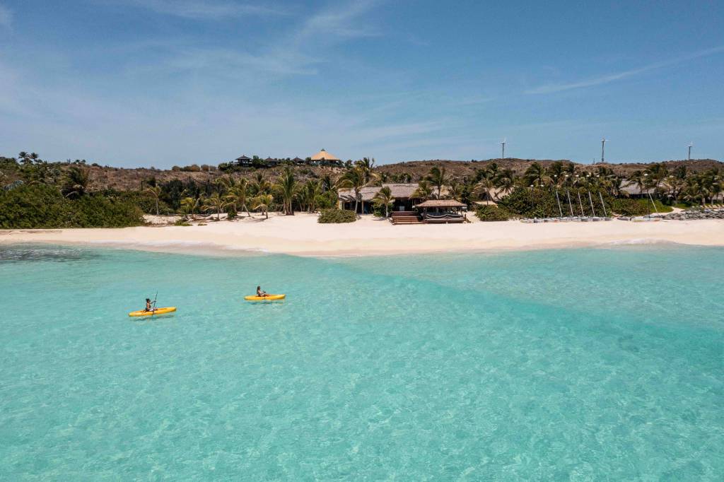 Duas pessoas em caiaques amarelos remam em águas cristalinas de uma praia paradisíaca, com areia branca e coqueiros. Ao fundo, casas com telhados de palha e turbinas eólicas em uma colina sob céu azul
