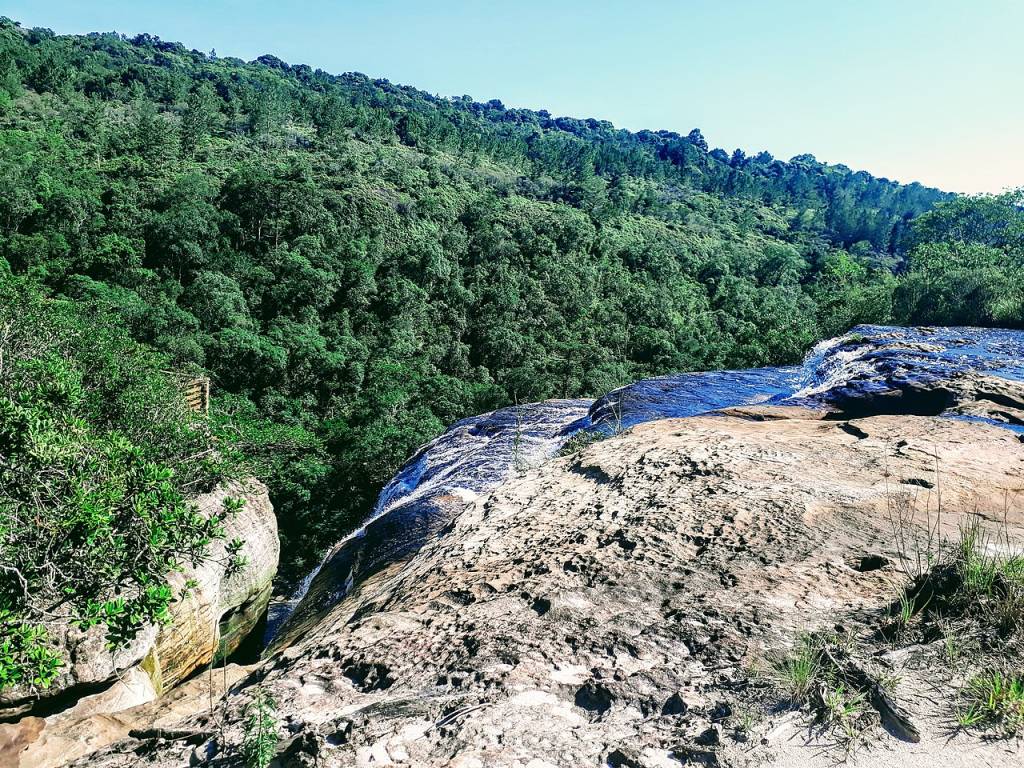 Vista de cima de uma cachoeira, com água azul escura fluindo sobre rochas claras e irregulares. Ao fundo, uma vasta floresta verde densa cobre uma montanha sob um céu azul claro. À esquerda, arbustos verdes e rochas grandes complementam a paisagem natural