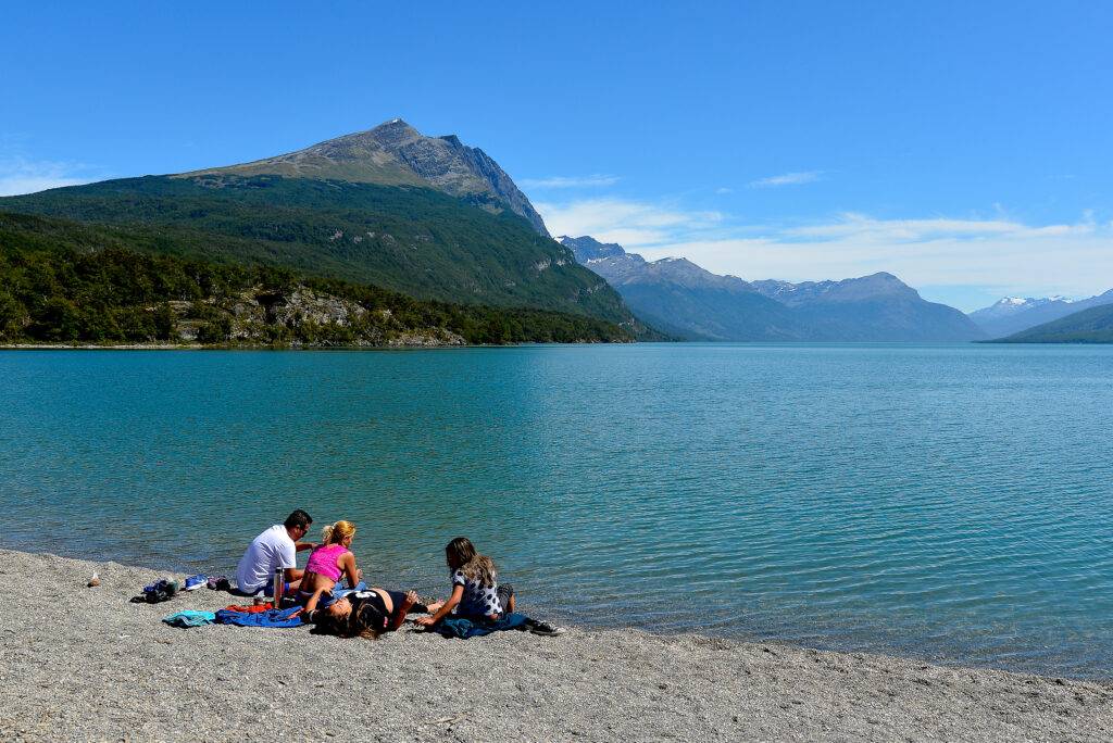 Quatro pessoas sentadas em uma praia de pedras, à beira de um lago azul-turquesa. Ao fundo, montanhas verdes e picos nevados sob céu azul