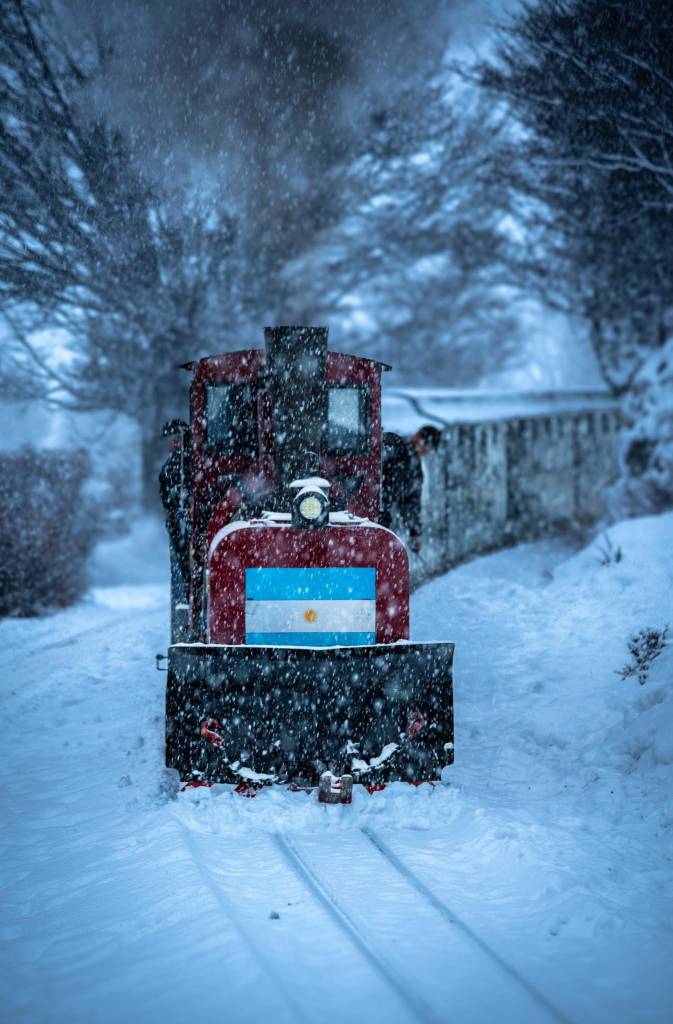 Locomotiva vermelha com a bandeira da Argentina na frente, coberta de neve, em trilhos nevados. Flocos de neve caem intensamente, com árvores ao fundo e um homem na cabine.