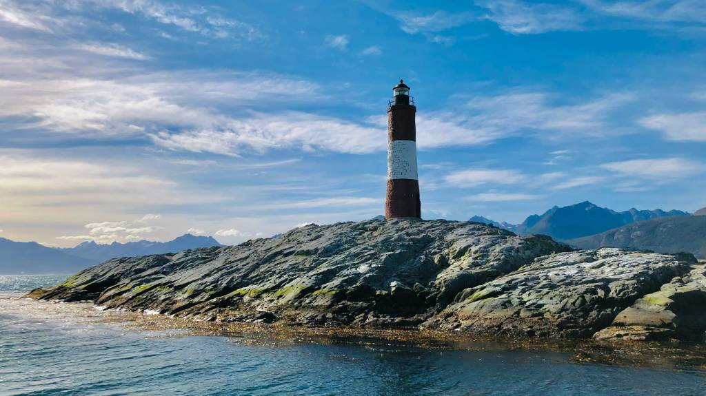 Farol Les Éclaireurs, vermelho e branco, sobre uma ilha rochosa no Canal de Beagle, com montanhas ao fundo e céu azul com nuvens esparsas