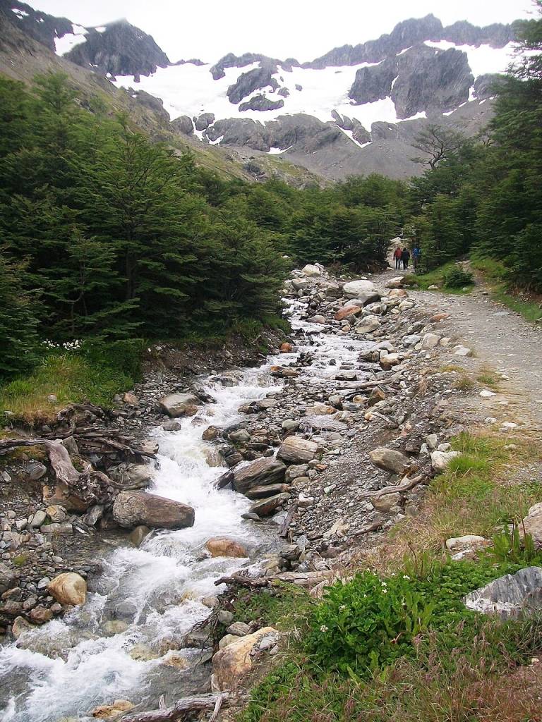 Riacho rochoso com água corrente e borbulhante, ladeado por árvores verdes e uma trilha de terra onde duas pessoas caminham. Ao fundo, montanhas cinzentas com picos nevados sob um céu nublado. Foto sob licença CC BY-SA 3.0
