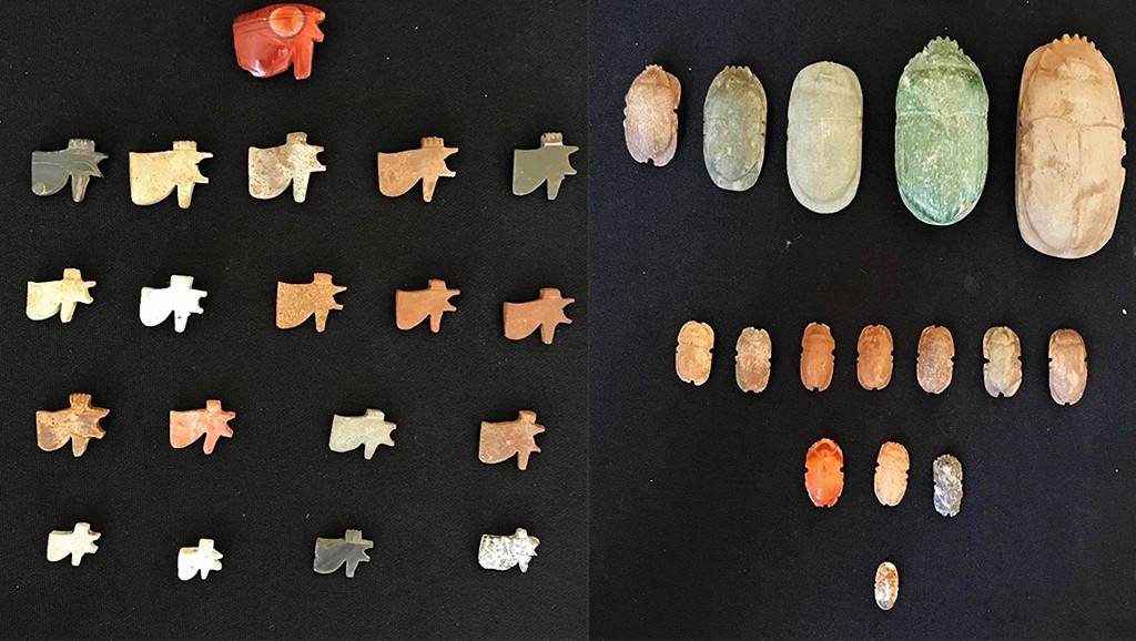 Coleção de 28 amuletos egípcios antigos sobre fundo preto. À esquerda, 19 amuletos em forma de machado, um vermelho e os demais em tons de bege, marrom e cinza. À direita, 9 amuletos em forma de escaravelho, variando em tamanho e cor, de verde a marrom claro, com um pequeno escaravelho branco na base