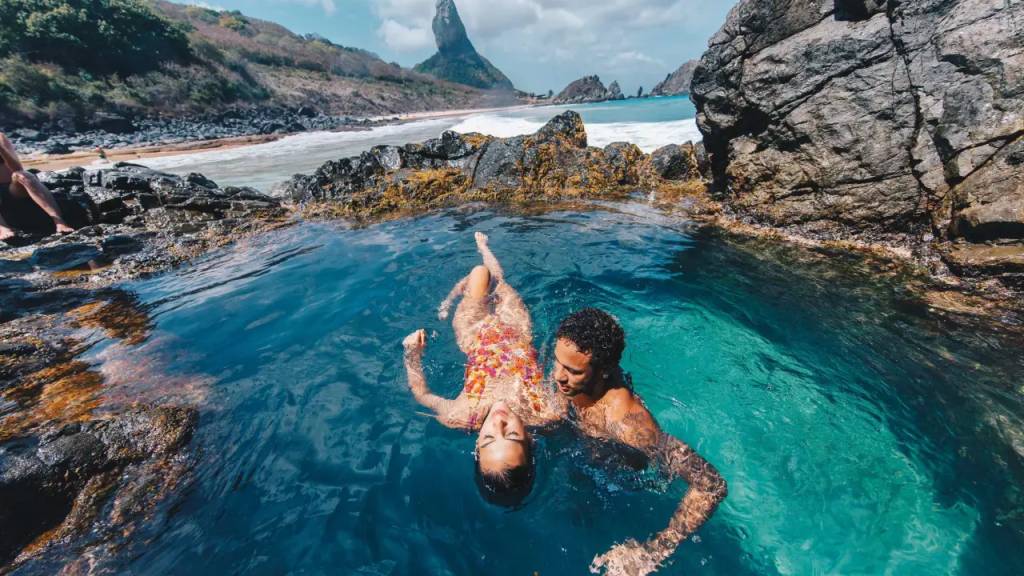 Casal jovem flutuando em piscina natural de águas claras, cercada por rochas escuras. Ao fundo, praia com ondas, vegetação e o Morro do Pico em Fernando de Noronha