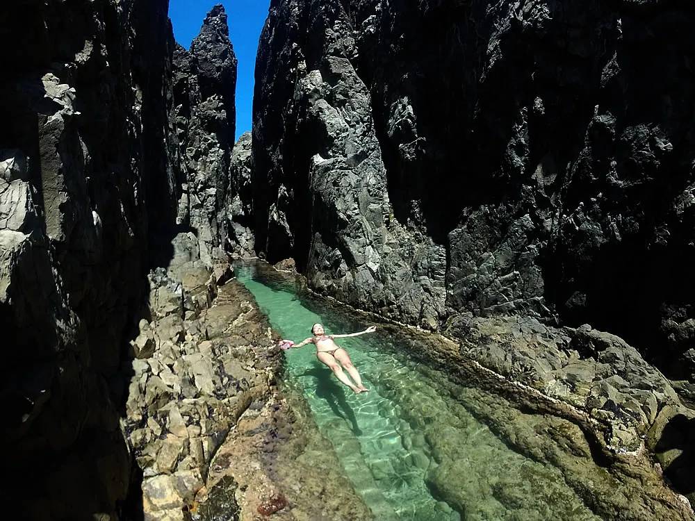 Mulher de biquíni flutuando de costas em uma piscina natural de água cristalina, cercada por paredões rochosos íngremes sob céu azul