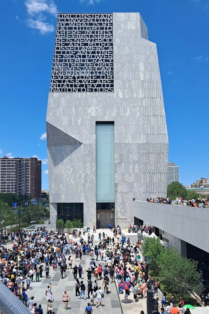 Edifício moderno de mármore cinza com fachada preta exibindo texto branco, UNCONSTRAIN CONVENTION BY WHAT IS RE THOUGHT TO WHERE IN THIS ARE FIRST STEPS RE IS NEW GROU RE ARE MORE OSSED AMERICA CL OF ANY ONE IF MOST POWER R DEMOCRACY WE THE PEOPLE OME YES WE CAN VNED BY NO ONE EVERYONE OF IS TASK WE ARE NUALLY TRY TO NATION OF OURS. Uma multidão de pessoas caminha em frente ao prédio e em uma passarela elevada à direita, sob céu azul com nuvens esparsas