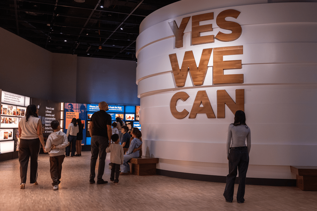Pessoas visitam um museu com paredes brancas e telas azuis exibindo fotos e textos. Uma parede curva exibe as palavras YES WE CAN em letras grandes de madeira. Uma mulher de costas observa a parede, enquanto famílias caminham e outras pessoas sentam em bancos de madeira, apreciando as exposições