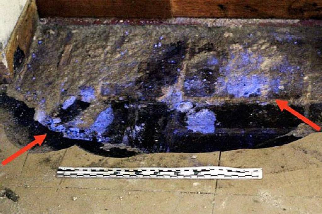 Manchas azul-violeta fluorescentes, indicando vestígios de sangue, em um piso de concreto danificado, com duas setas vermelhas apontando para as áreas de maior concentração. Uma régua de escala branca e preta está no chão, abaixo das manchas