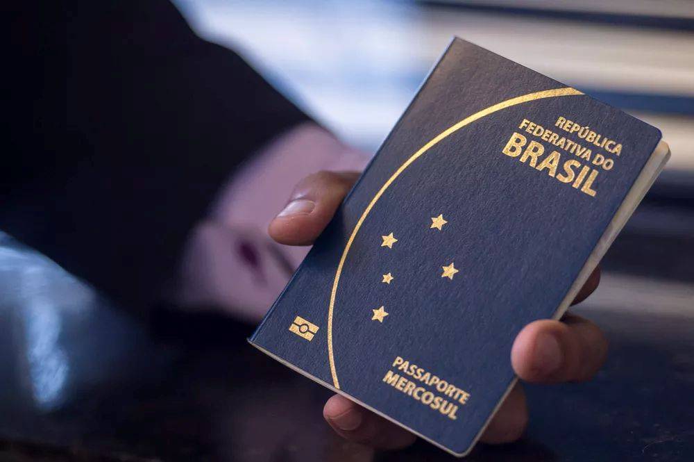 Mão de uma pessoa segurando um passaporte brasileiro azul-marinho com detalhes dourados, incluindo as palavras REPÚBLICA FEDERATIVA DO BRASIL e PASSAPORTE MERCOSUL