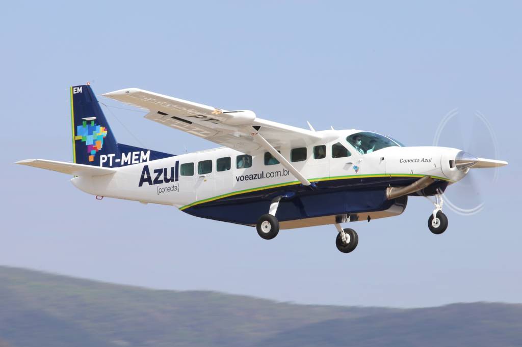 Avião monomotor branco e azul da Azul Conecta, modelo Cessna Caravan, com hélice girando, voando baixo contra um céu azul claro. A cauda tem um mapa do Brasil colorido em pixels e a matrícula PT-MEM. O trem de pouso está abaixado
