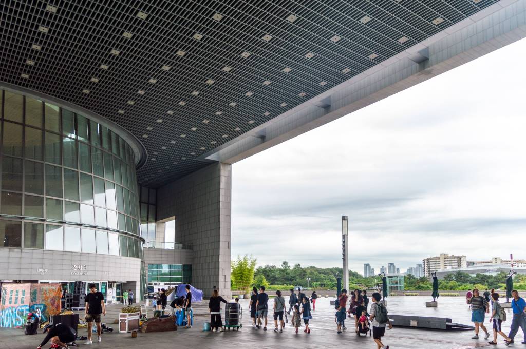 Pessoas caminham em uma praça ampla com um edifício moderno de fachada curva e vidro à esquerda, sob um teto de treliça metálica. Ao fundo, árvores e prédios urbanos sob um céu nublado