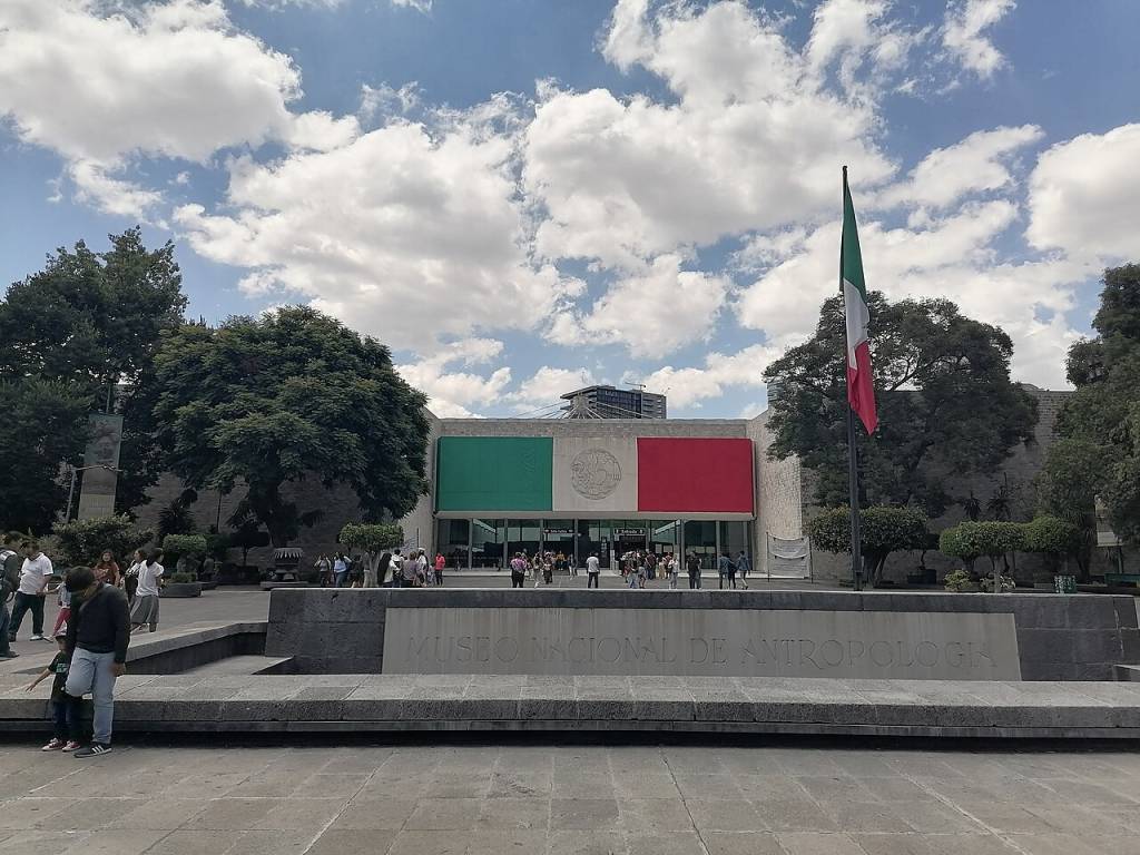 Fachada do Museu Nacional de Antropologia, México, com painéis verde e vermelho e um brasão central. Uma bandeira mexicana tremula à direita. Pessoas caminham na praça em frente, sob um céu azul com nuvens brancas. O nome MUSEO NACIONAL DE ANTROPOLOGIA está gravado em uma estrutura de pedra em primeiro plano