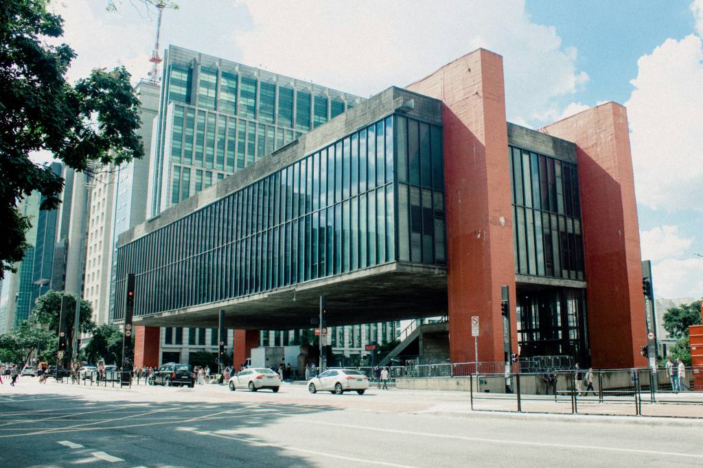 Vista externa do MASP em São Paulo, um edifício de concreto suspenso por quatro pilares vermelhos, com fachada de vidro e telhado plano. Abaixo, carros e pedestres transitam na Avenida Paulista, sob céu azul com nuvens esparsas