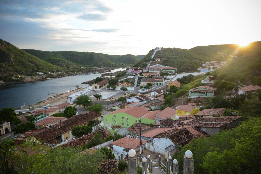 Vista aérea de Piranhas, Alagoas, com casas coloridas e telhados de barro, margeando o rio São Francisco. O sol se põe à direita, iluminando as montanhas verdes e o céu nublado. Uma escadaria branca sobe a encosta da montanha.