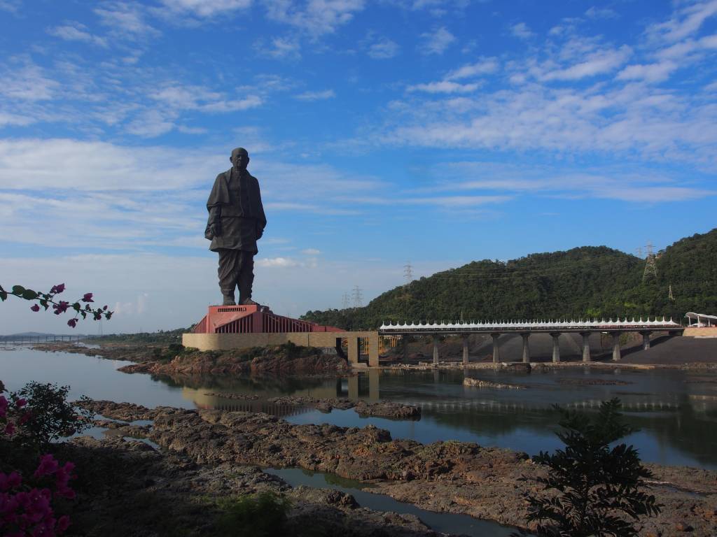 Estátua da Unidade, uma figura masculina imponente, em um pedestal vermelho, à beira de um rio com margens rochosas. Ao fundo, uma ponte longa e montanhas verdes sob um céu azul com nuvens esparsas. Foto sob licença CC BY-SA 4.0
