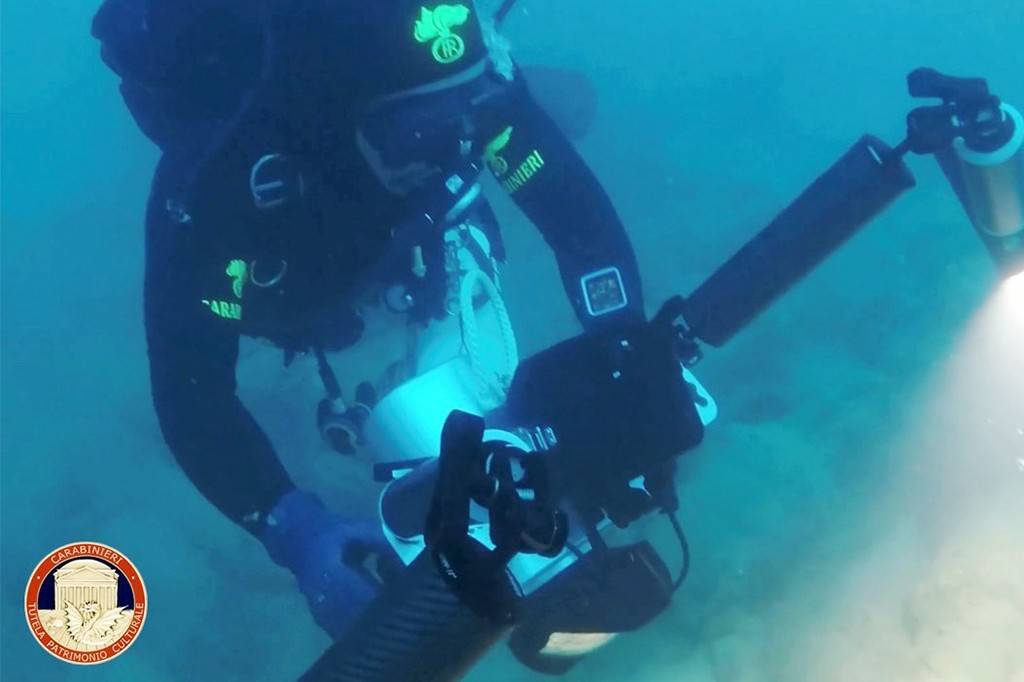 Mergulhador com roupa de neoprene preta e luvas azuis, usando máscara e equipamento de mergulho, segura uma câmera subaquática com luz acesa, iluminando a água turva. O emblema dos Carabinieri aparece no canto inferior esquerdo