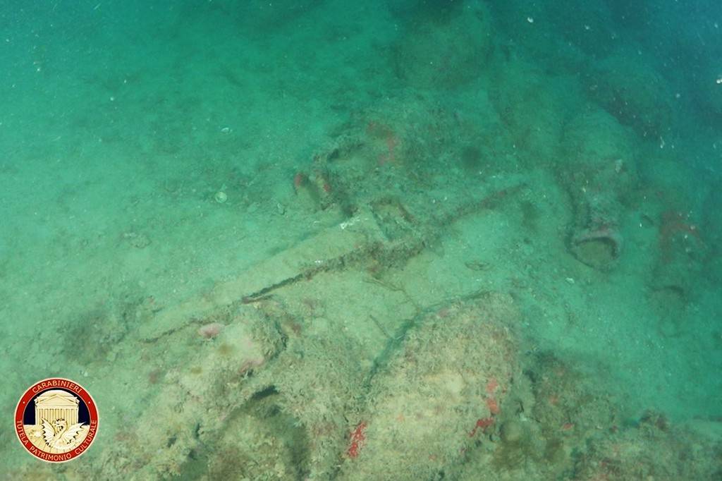 Fundo do mar turquesa com areia e rochas cobertas por algas, revelando fragmentos de ânforas e outros artefatos antigos submersos. No canto inferior esquerdo, o logo dos Carabinieri.