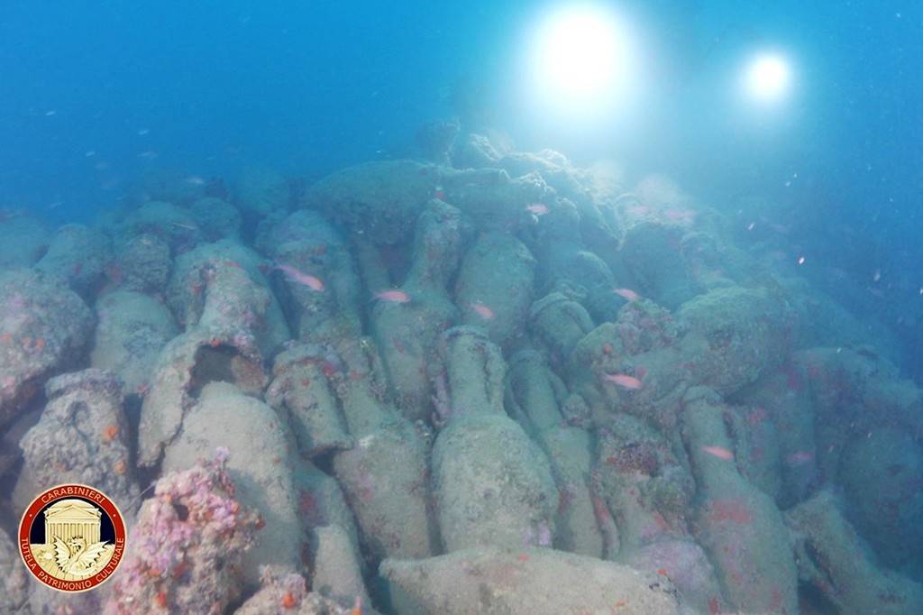 Várias ânforas antigas cobertas por incrustações marinhas e corais, repousando no fundo do mar. Pequenos peixes rosados nadam entre elas. A luz do sol penetra a água azul-escura, iluminando o cenário. No canto inferior esquerdo, um selo vermelho com a inscrição Carabinieri Tutela Patrimonio Culturale