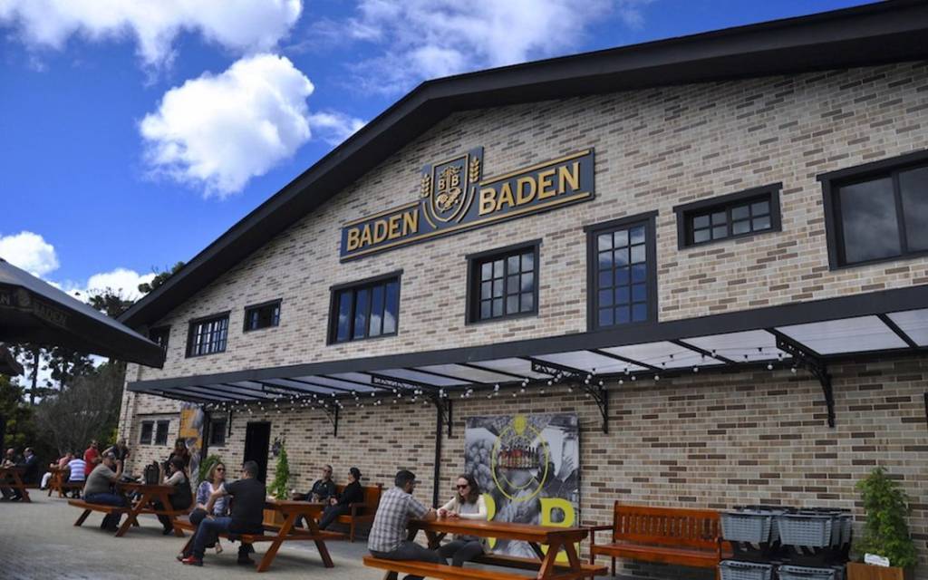 Fachada de tijolos da cervejaria Baden Baden com letreiro dourado e preto, sob céu azul com nuvens. Pessoas sentadas em mesas de madeira ao ar livre, com guarda-sóis e um toldo transparente com luzes penduradas.