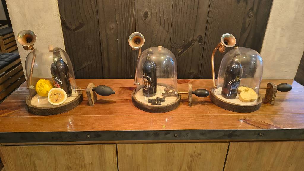 Três garrafas de cerveja Baden Baden em cúpulas de vidro, com elementos decorativos como maracujá, chocolates e biscoitos, sobre um balcão de madeira rústica