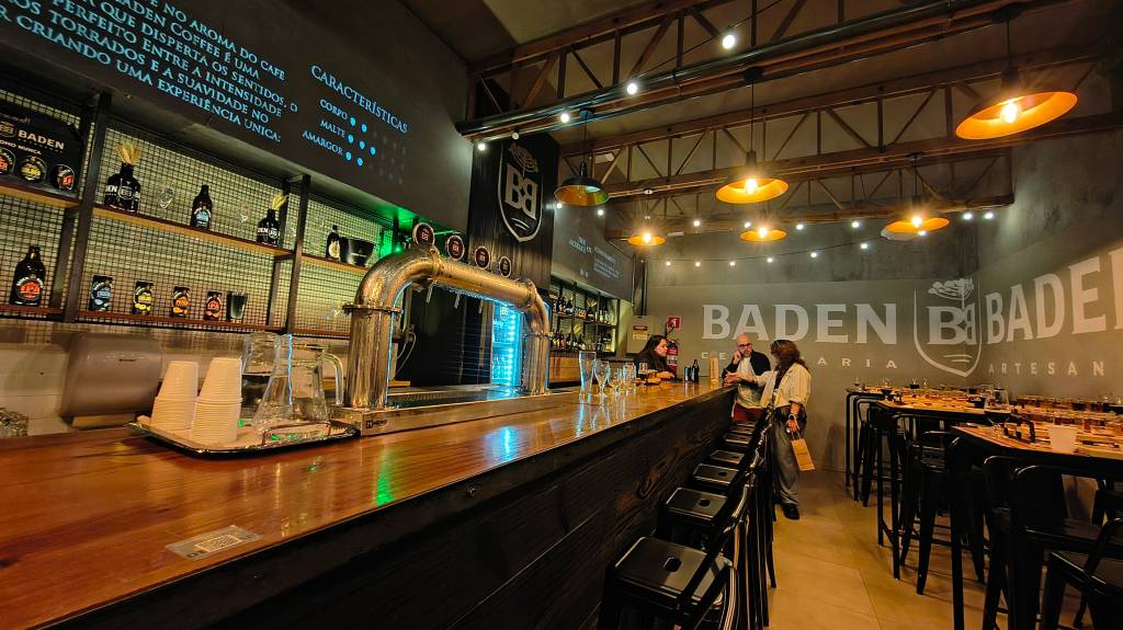 Interior de uma cervejaria Baden Baden com balcão de madeira, torneiras de chope e prateleiras com garrafas. No fundo, pessoas conversam em mesas altas com bancos, sob iluminação quente e letreiros da marca na parede