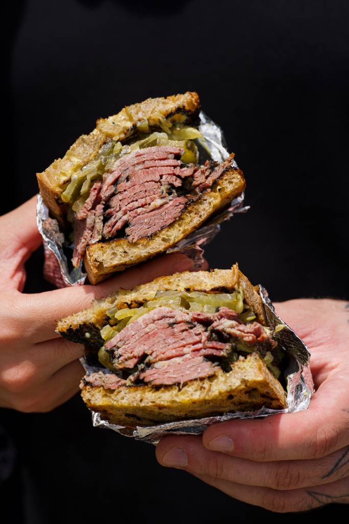 Mãos seguram um sanduíche de pastrami cortado ao meio, revelando fatias de carne rosada, picles e cebola caramelizada entre fatias de pão tostado, parcialmente embrulhado em papel alumínio