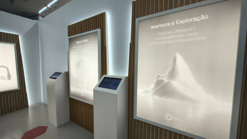 Exposição com três painéis luminosos e dois totens interativos em um ambiente moderno. O painel da direita exibe Aventura e Exploração com uma montanha branca, o central Impacto Global com uma árvore, e o da esquerda uma imagem parcial de um braço. Os totens brancos têm telas azuis.