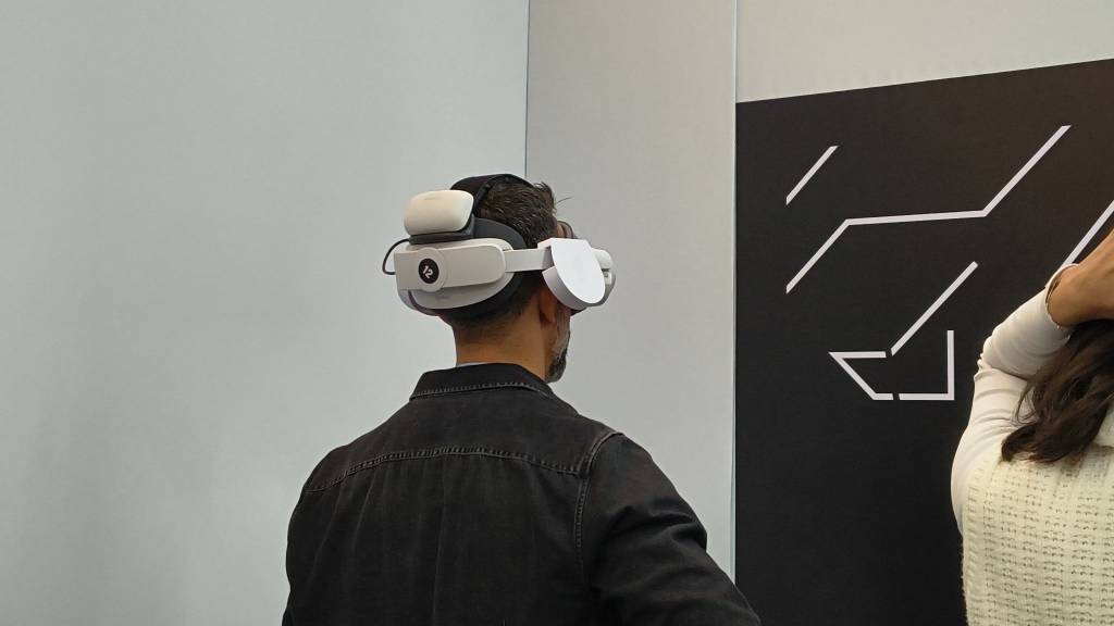 Homem de costas, vestindo jaqueta jeans escura, usando óculos de realidade virtual brancos e cinzas, com um fone de ouvido branco na parte de trás da cabeça. Ele está em um ambiente interno com paredes claras e um painel preto com linhas brancas abstratas. Uma mulher de blusa clara aparece parcialmente à direita