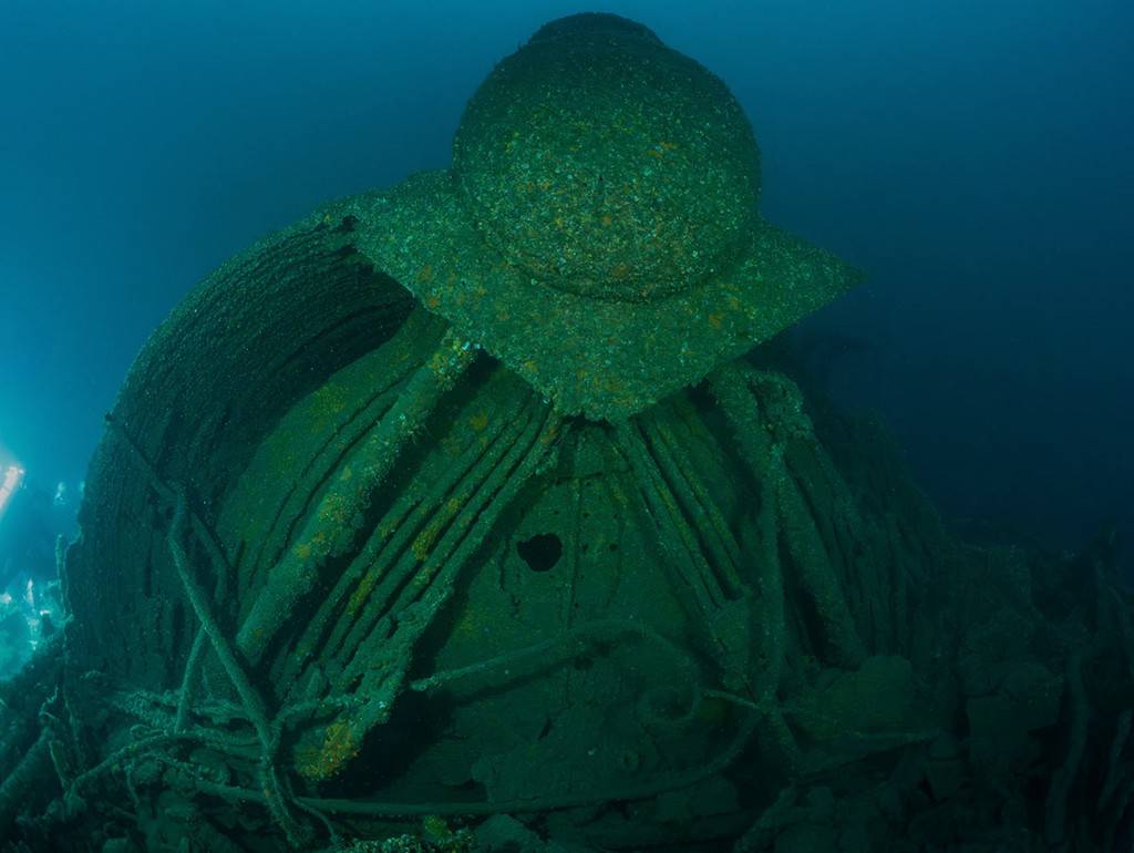 Parte de um naufrágio submerso, coberto por algas verdes e incrustações, com detalhes de metal enferrujado e estruturas retorcidas no fundo do mar azul escuro