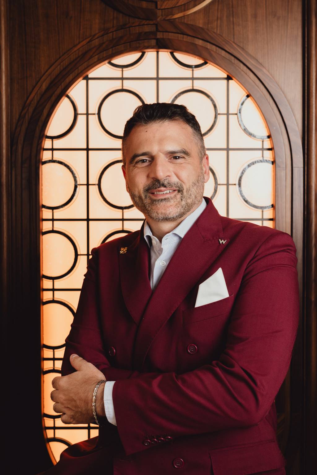 Homem de meia-idade, barba grisalha e sorriso gentil, veste um terno vinho com camisa listrada e lenço branco no bolso, braços cruzados. Ele está em pé, em frente a uma janela decorativa de madeira escura com detalhes circulares e retangulares, iluminada por trás