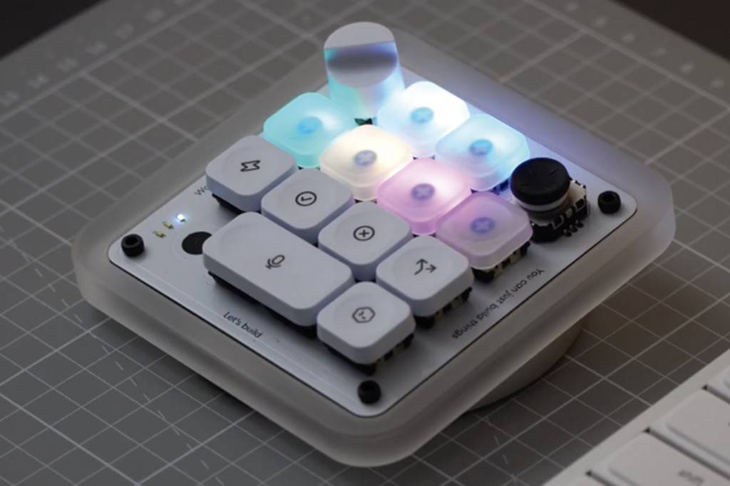 SI_490_Tech_Teclado_OpenAI Um teclado modular branco com teclas iluminadas em tons de azul, roxo e branco, sobre uma base quadriculada cinza. Algumas teclas têm ícones como microfone, sigma e setas. Há um joystick preto no canto superior direito e texto Lets build e You can just build things na base