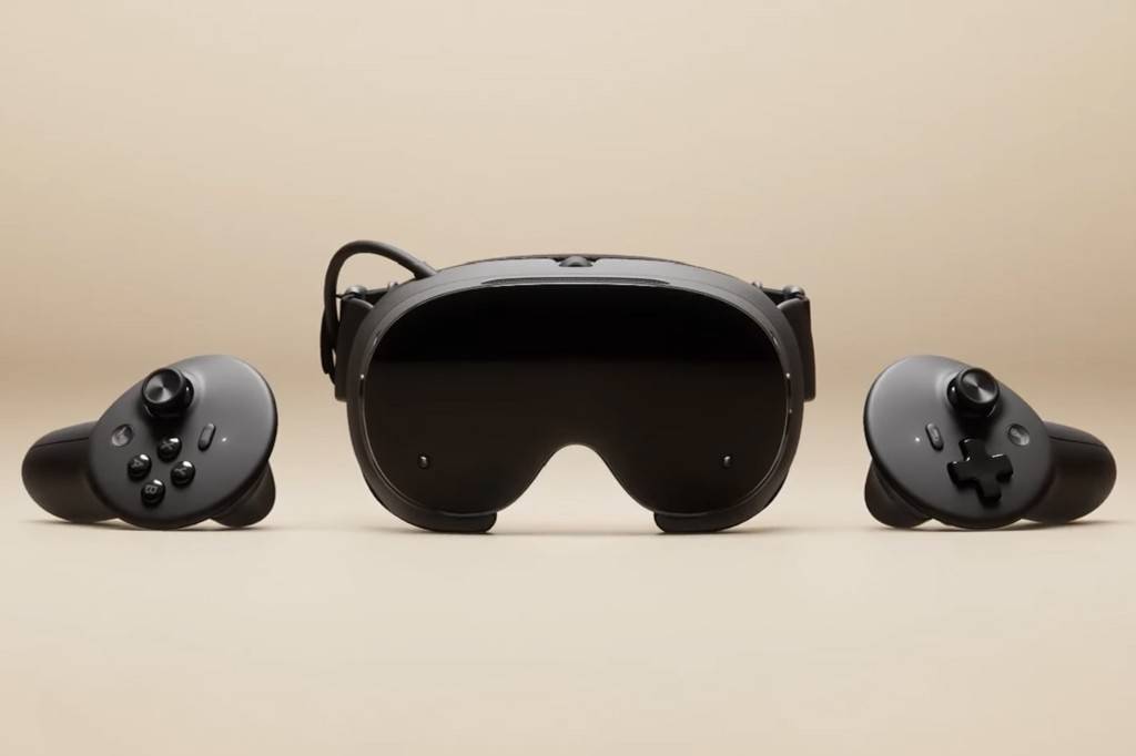 SI_490_Tech_Capacete_Valve Um óculos de realidade virtual preto com lentes escuras e dois controles pretos, um de cada lado, sobre um fundo bege.