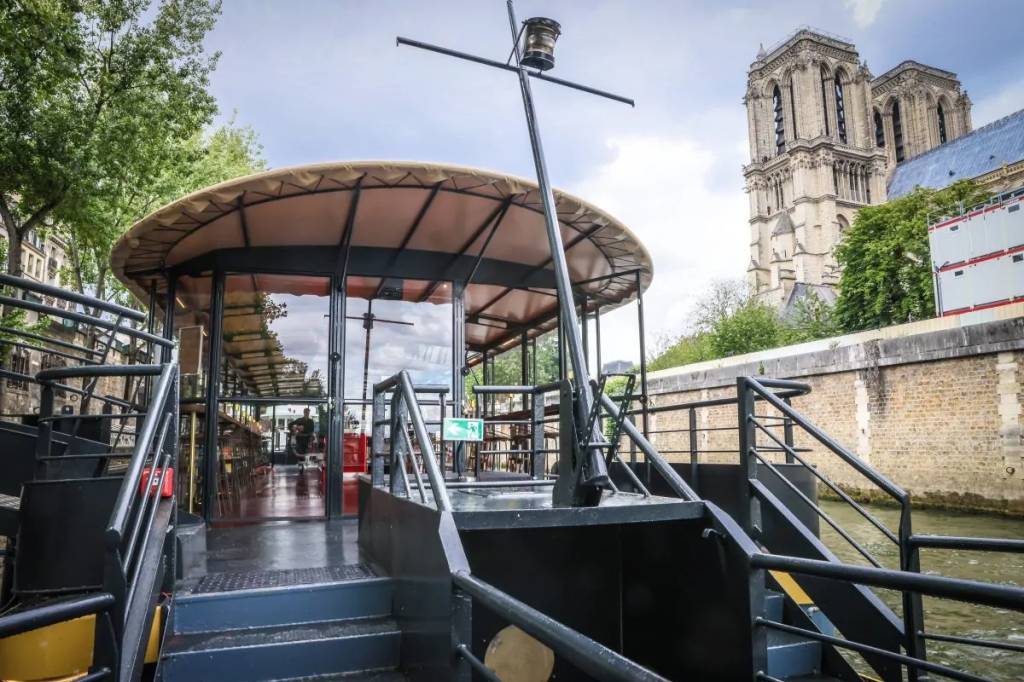 Vista da proa de um barco turístico no rio Sena, com a Catedral de Notre-Dame ao fundo. O barco tem um teto de lona bege e janelas de vidro, com degraus e corrimãos pretos. A catedral, de pedra clara, exibe suas duas torres góticas sob um céu nublado. Árvores verdes margeiam o rio, e um muro de pedra clara delimita a margem.
