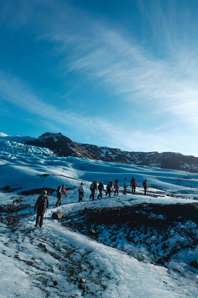 Grupo de pessoas caminhando em fila sobre uma geleira azul-clara, com montanhas rochosas e um céu azul vibrante com nuvens finas ao fundo