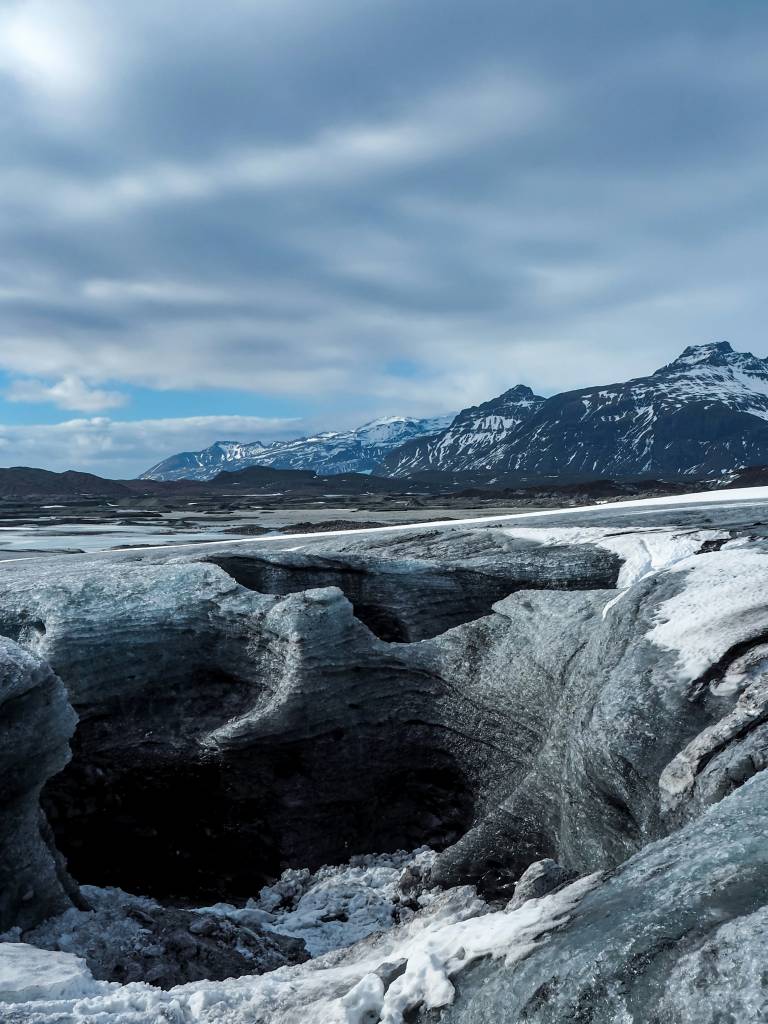 Geleira com grandes fendas e gelo escuro no primeiro plano, montanhas nevadas ao fundo e céu nublado
