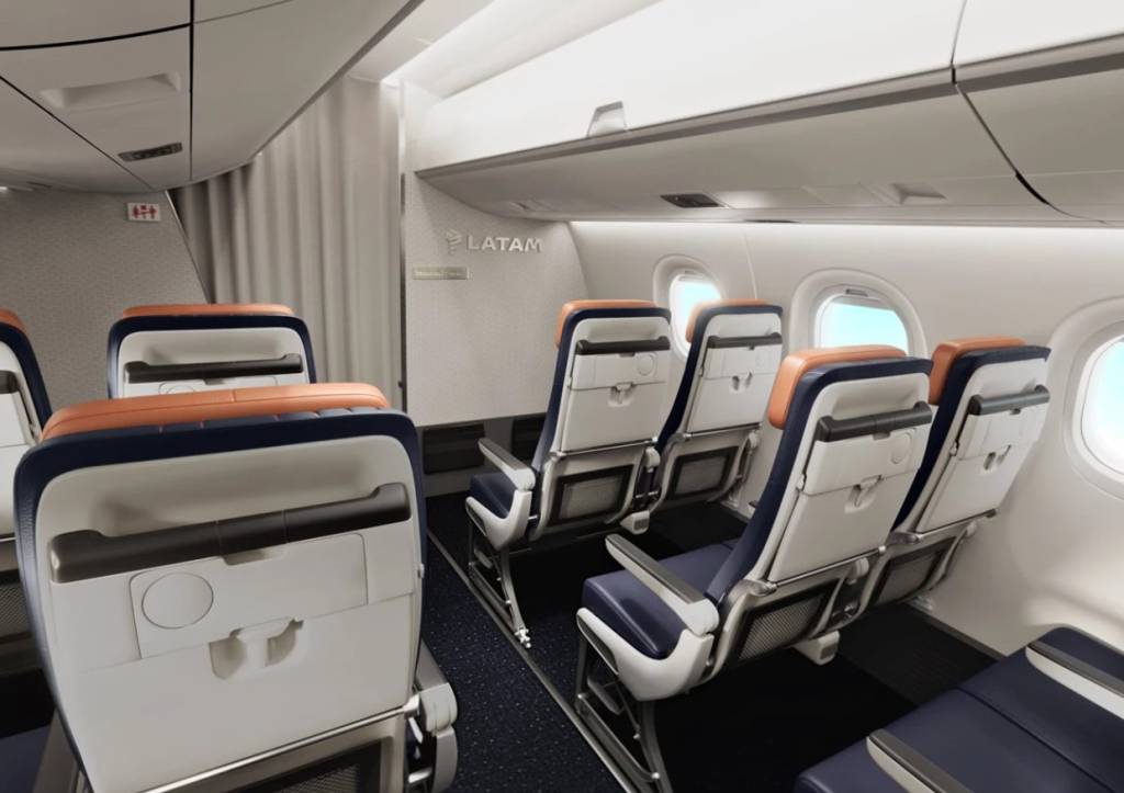 Interior de um avião LATAM com fileiras de assentos vazios. Os assentos são azuis escuros e brancos, com detalhes em laranja nos encostos de cabeça. As janelas ovais mostram um céu azul claro. O logotipo da LATAM está visível na parede frontal da cabine