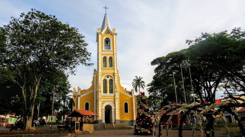 Igreja amarela com torre alta e telhado cinza pontiagudo, ladeada por árvores frondosas e uma palmeira. À direita, uma árvore de Natal decorada com laço vermelho e enfeites dourados. No primeiro plano, uma barraca de madeira com telhado vermelho e decorações natalinas. O céu está claro com algumas nuvens