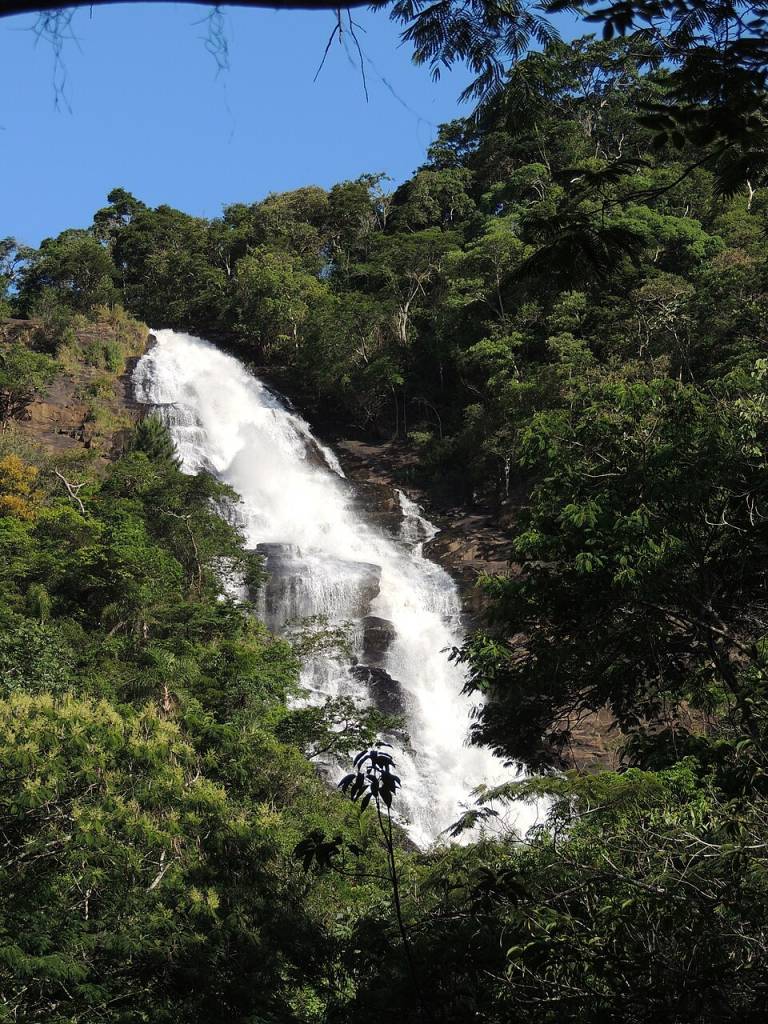 Cachoeira volumosa descendo por rochas escuras, cercada por densa vegetação verde sob um céu azul claro