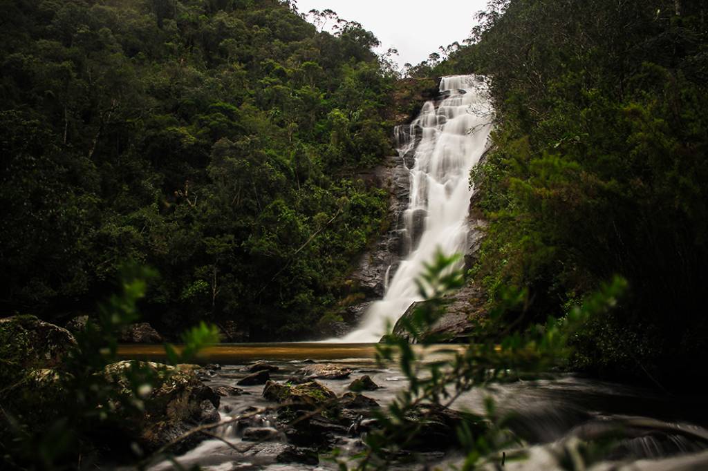 Cachoeira de múltiplos degraus com água branca e embaçada, caindo sobre rochas escuras, cercada por densa floresta verde. Um rio marrom flui na base, com pedras e folhagens em primeiro plano, sob um céu nublado. Foto sob licença CC BY-SA 3.0