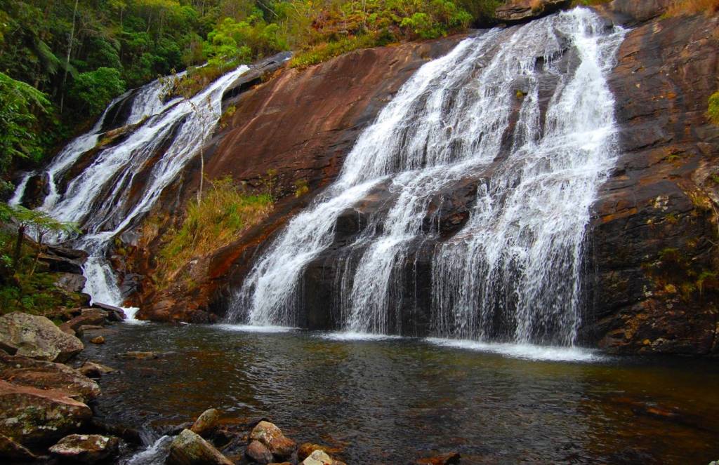 Cachoeira com várias quedas dágua descendo por uma rocha marrom-avermelhada, cercada por vegetação verde exuberante e um rio escuro com pedras no primeiro plano