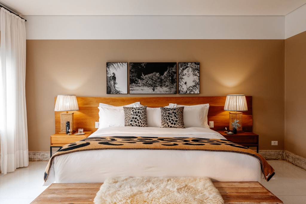 Quarto com cama de casal, lençóis brancos, duas almofadas de estampa de onça e manta marrom com manchas pretas. Duas mesas de cabeceira de madeira com abajures acesos e três quadros em preto e branco na parede bege. Um banco de madeira com tapete felpudo claro está aos pés da cama. Cortina branca na lateral esquerda