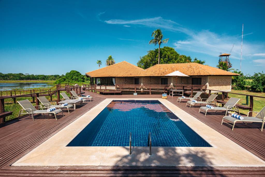Piscina retangular azul em deck de madeira, com espreguiçadeiras e toalhas listradas. Ao fundo, casas de telhado de barro, vegetação exuberante e um lago sob céu azul claro