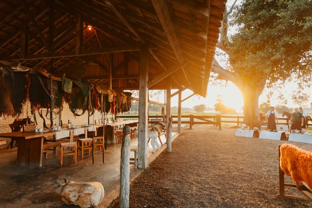 Rancho rústico com telhado de madeira, selas penduradas, mesas postas e bancos. Ao fundo, o sol se põe atrás de uma árvore grande, iluminando três homens com aventais e cavalos, preparando um evento ao ar livre